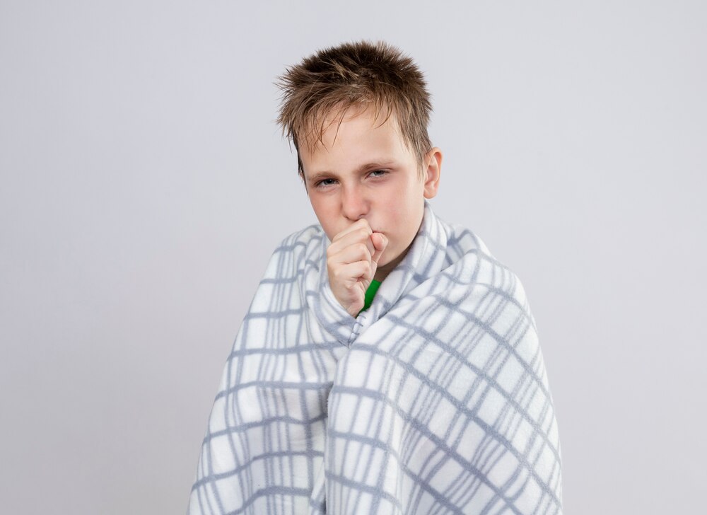 Ill little boywrapped in blanket feeling unwell coughing standing over white wall